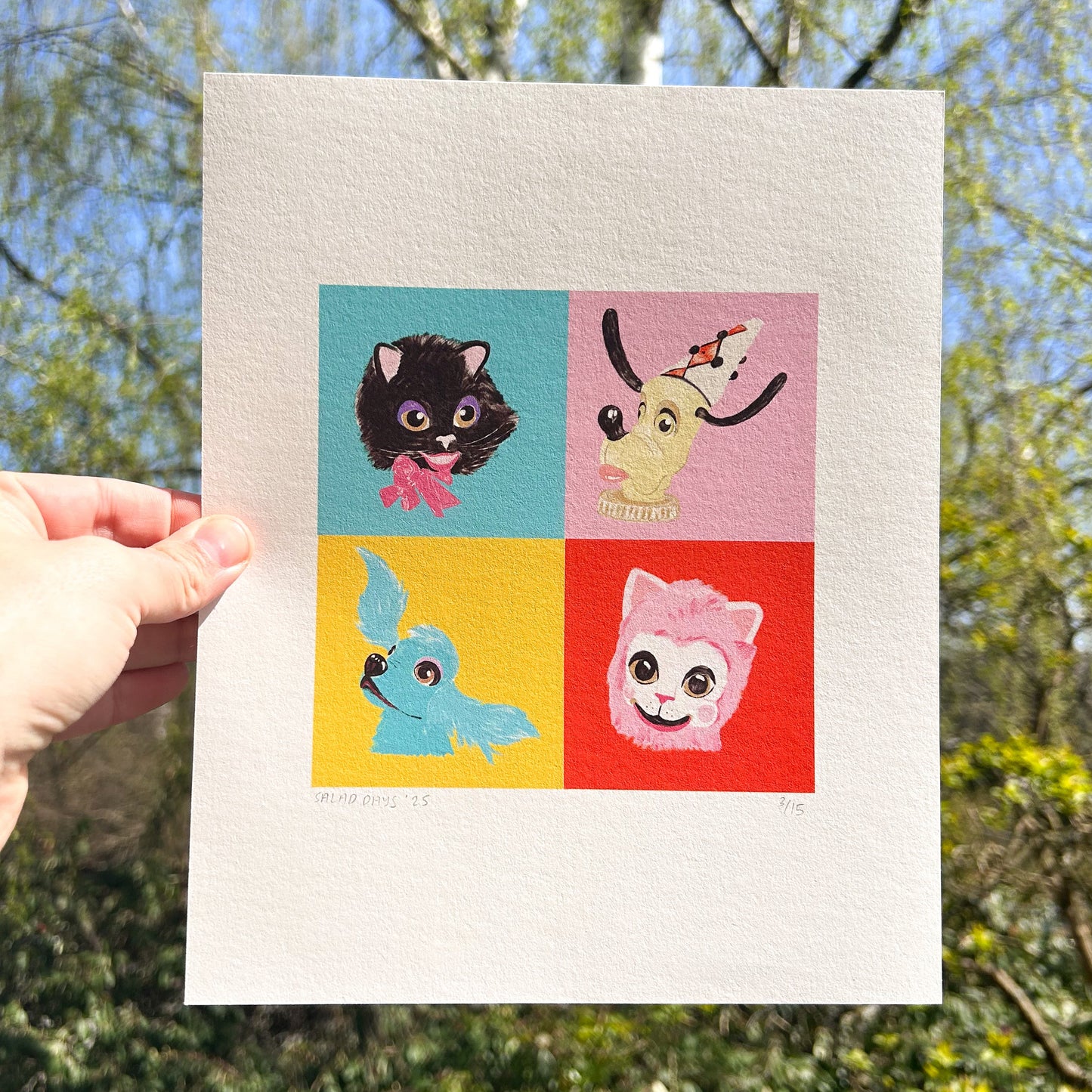 Four colorful illustrations of animal characters on a white card held outdoors with trees in the background.