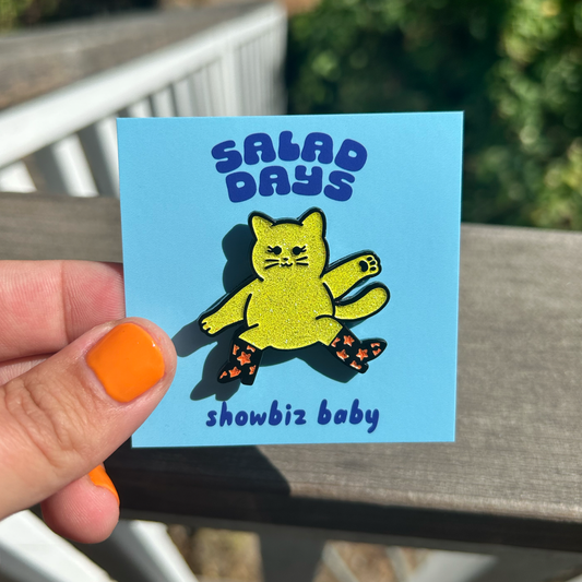 Showbiz Baby enamel pin featuring a glittery cat with boots and two it looks on the packaging